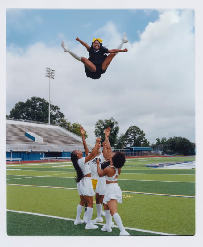Four cheerleaders in white uniforms lift another cheerleader in black high into the air on a football field, with empty bleachers and a cloudy sky in the background. The airborne cheerleader wears a yellow bow and smiles.