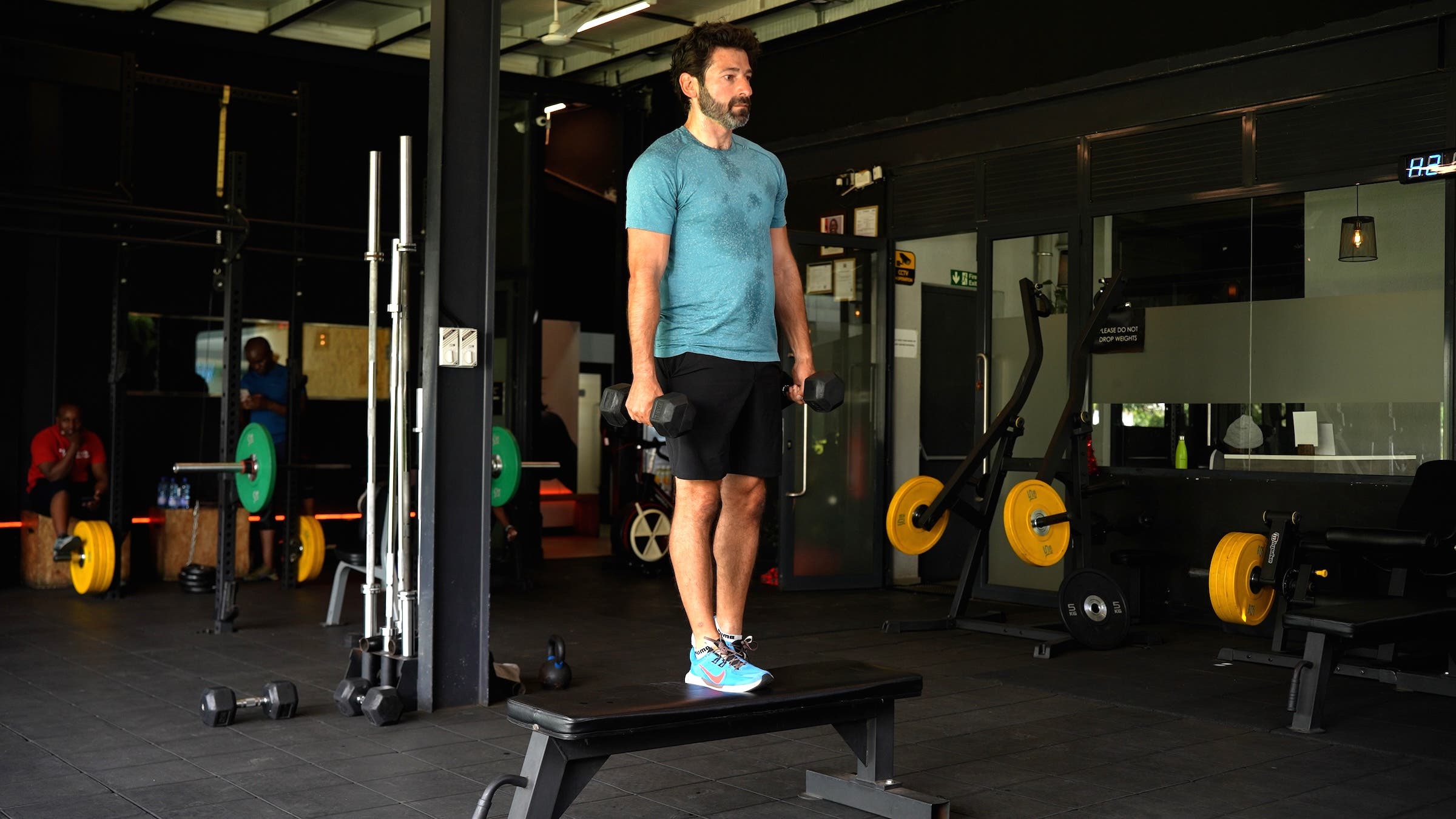 a man with dark hair wearing a blue shirt and black shorts demonstrates how to do a weighted step-up on a bench