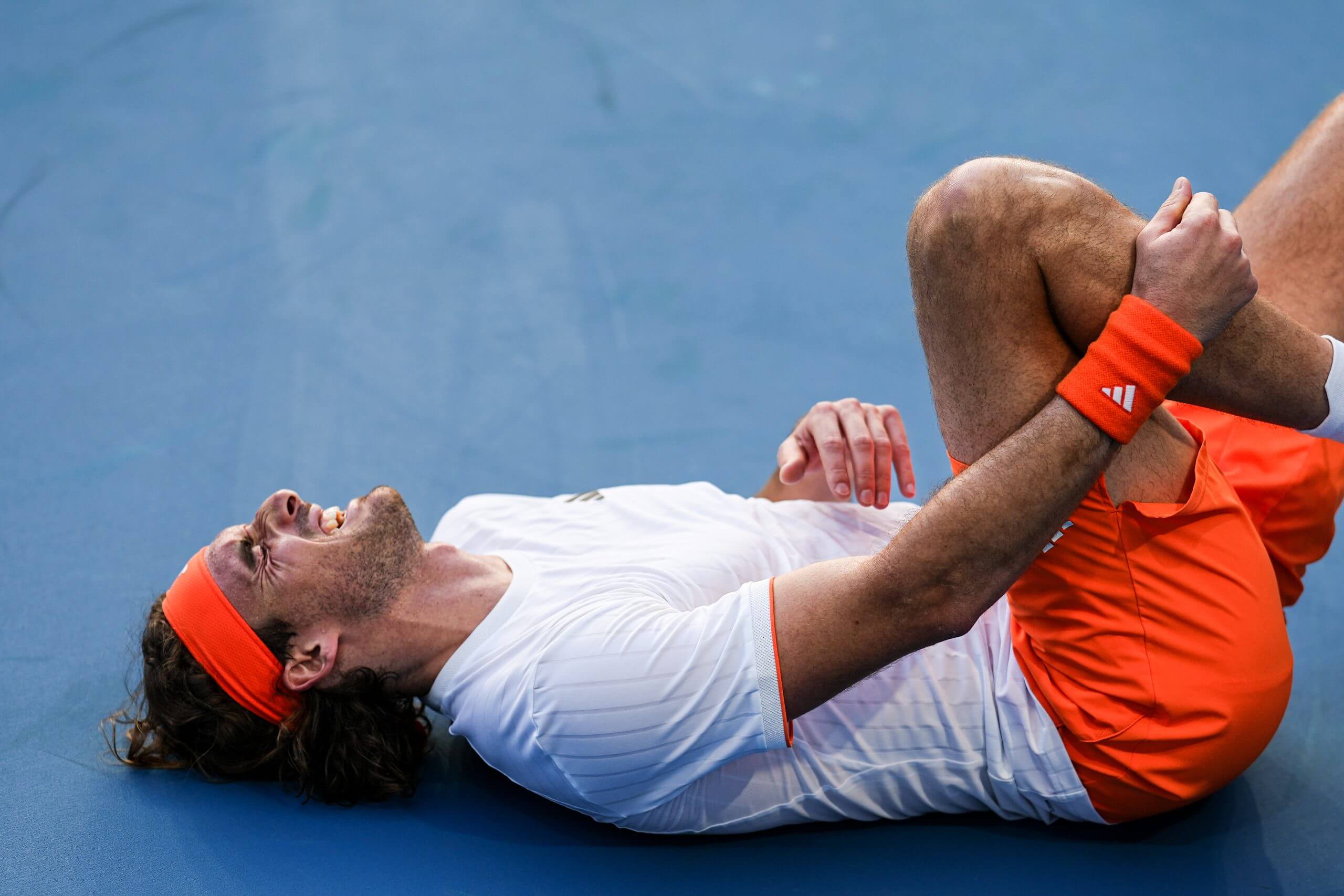 Stefanos Tsitisipas lies on a blue tennis court grasping his left leg just below the knee, grimacing in pain.