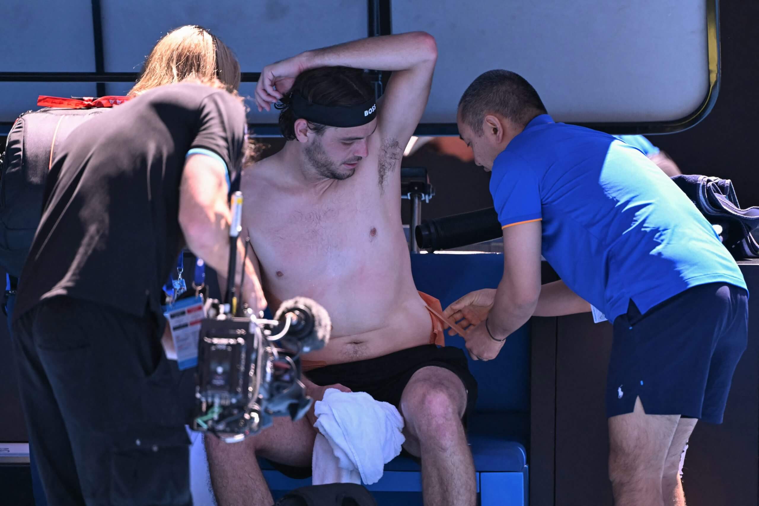 A trainer removes kinesthetic tape from Taylor Fritz's left side while he sits on a bench at the side of a tennis court.