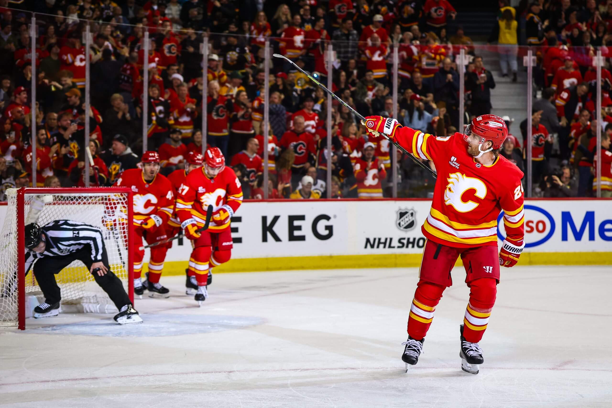 Blake Colman points his stick to a roaring crowd after scoring, with a ref grabbing the puck from the net and Flames teammates gathered nearby.