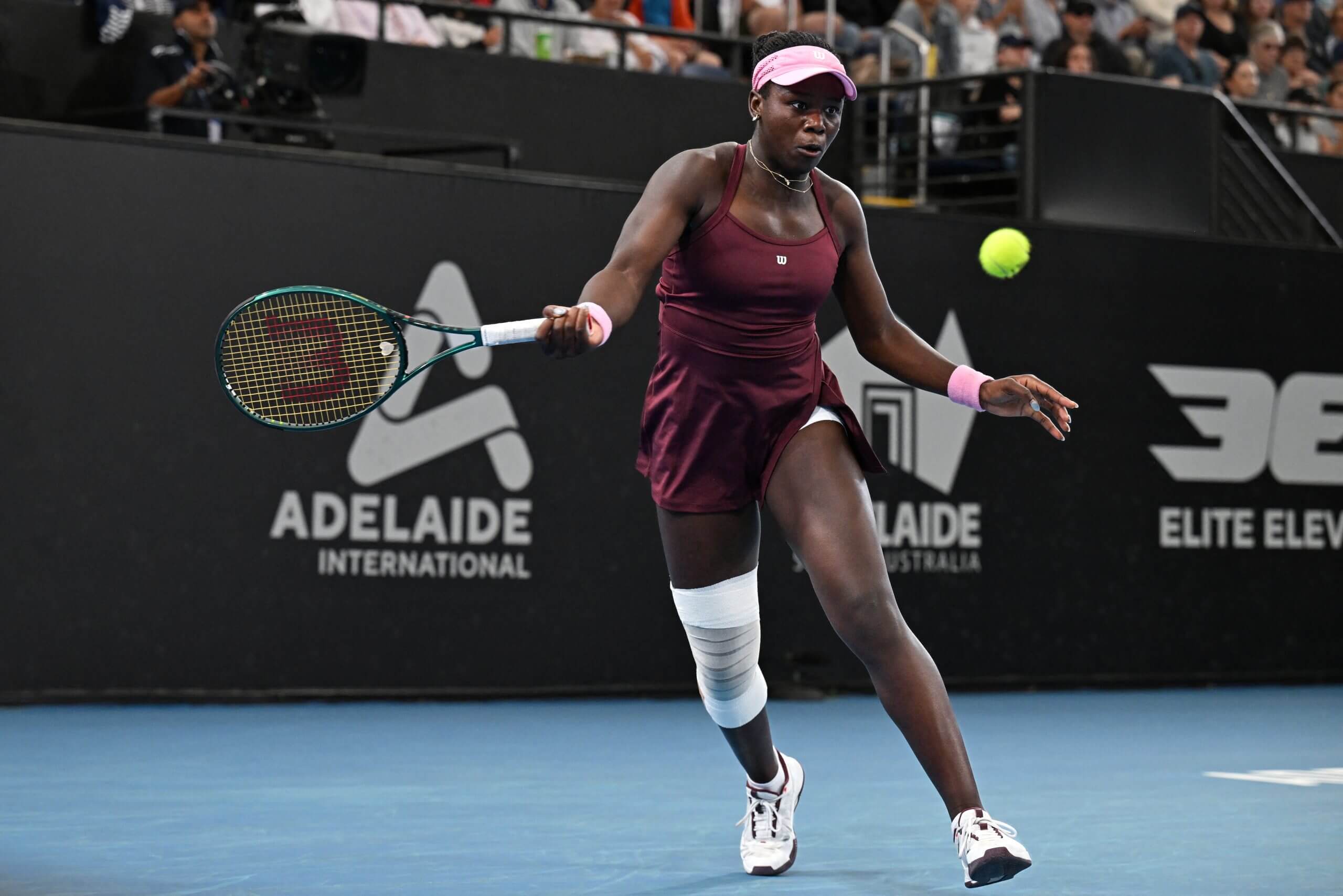 Victoria Mboko hits a forehand, wearing a burgundy tennis outfit with a white bandage on her right knee.