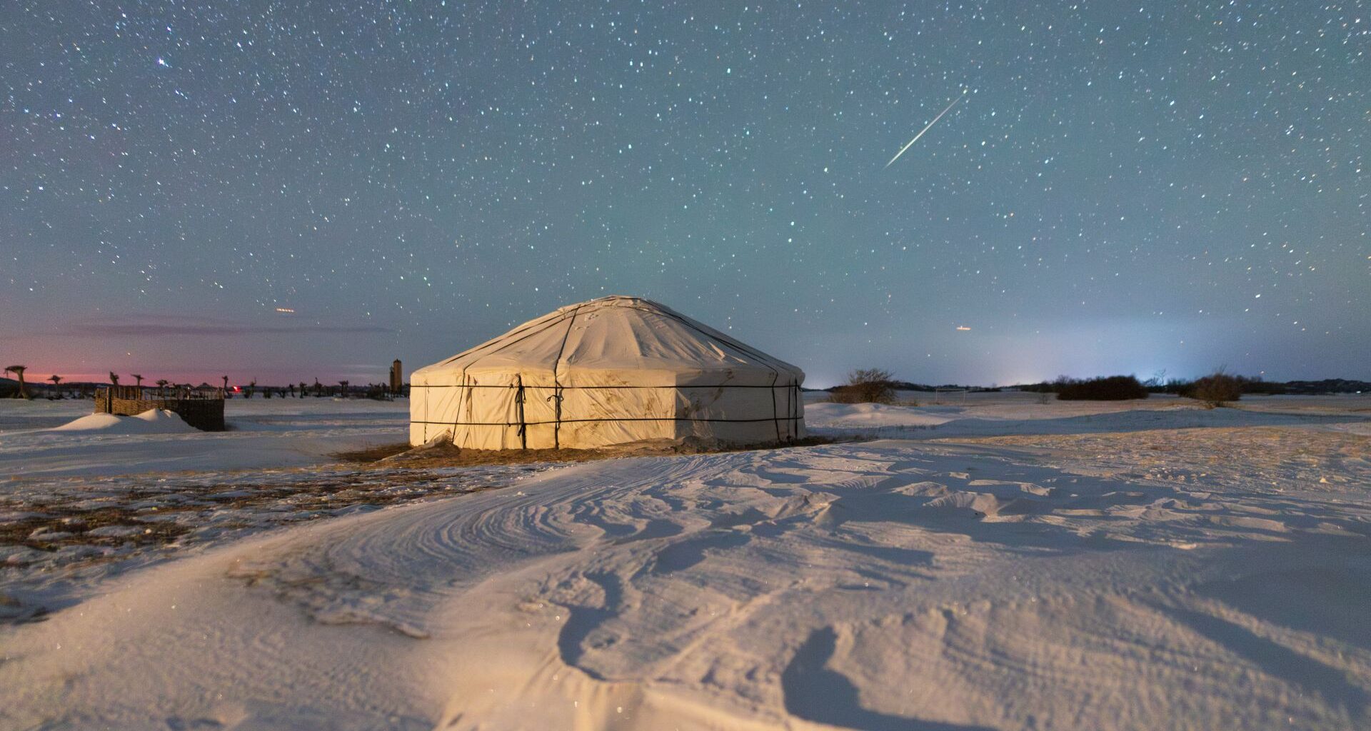 A meteor streaks throught the starry night sky above a fabric structure surrounded by snow.