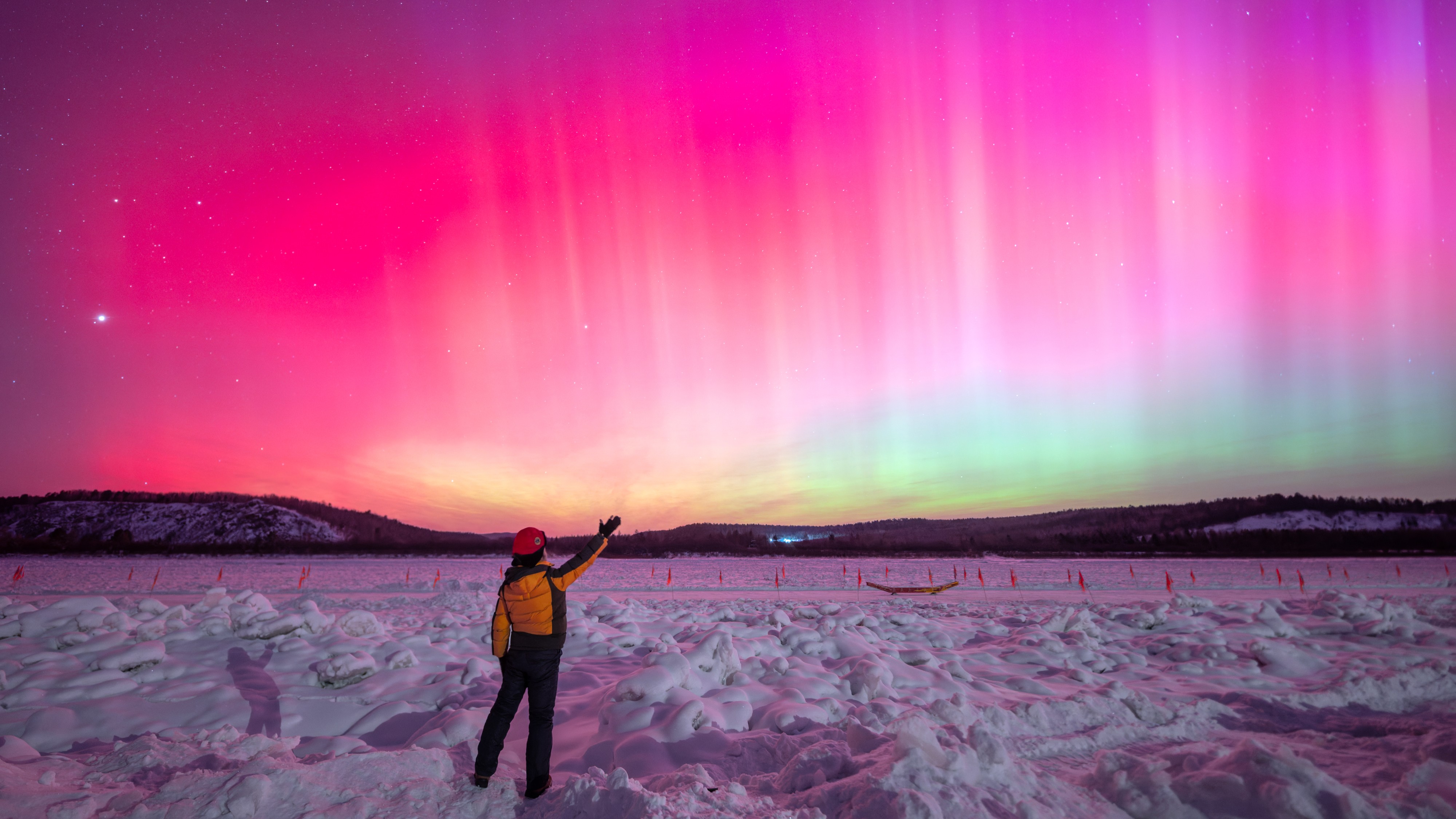 northern lights fill the sky with rich magenta colors and bright pillars of light. a person stands in the foreground of the image and reaches up to the sky with one hand.