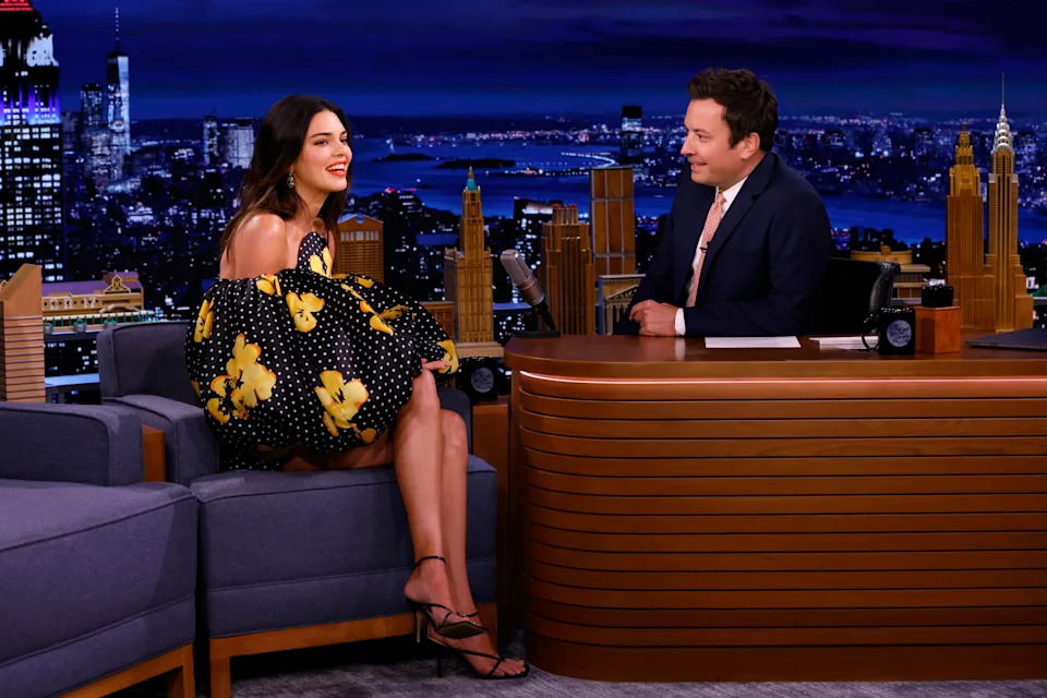 Kendall Jenner in a voluminous, off-shoulder dress with floral patterns is being interviewed on a talk show set at night