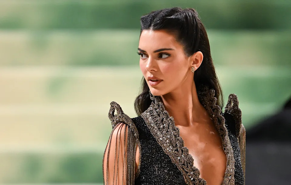 Kendall Jenner in ornate, sleeveless gown with delicate lace and fringe details at a formal event, gazing to the side with styled hair
