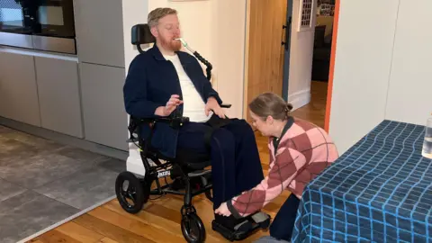 Michael Campbell is pictured in his wheelchair with his wife Naomi. He has a tube coming out of his mouth. His wife is bending down, securing his feet into straps of the wheelchair. MIchael is wearing dark clothing with a white T-shirt.
His wife has on a pink and purple jumper in a diamond pattern and dark trousers. Her hair is in a bun.