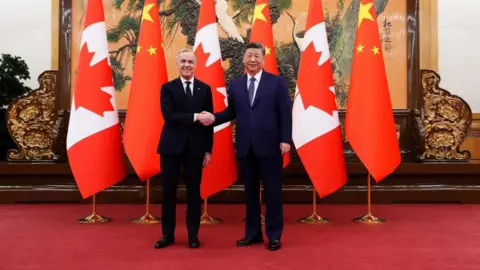 Reuters Canadian Prime Minister Mark Carney smiles as he shakes hands with President of China Xi Jinping at the Great Hall of the People in Beijing. Facing the camera, they stand on a red carpet in front of Canadian and Chinese flags. 