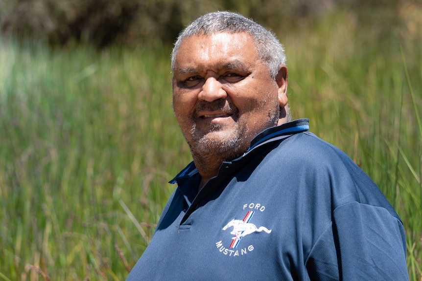 A smiling, middle-aged man with short, greying hair wears a branded polo shirt while standing in grassland.
