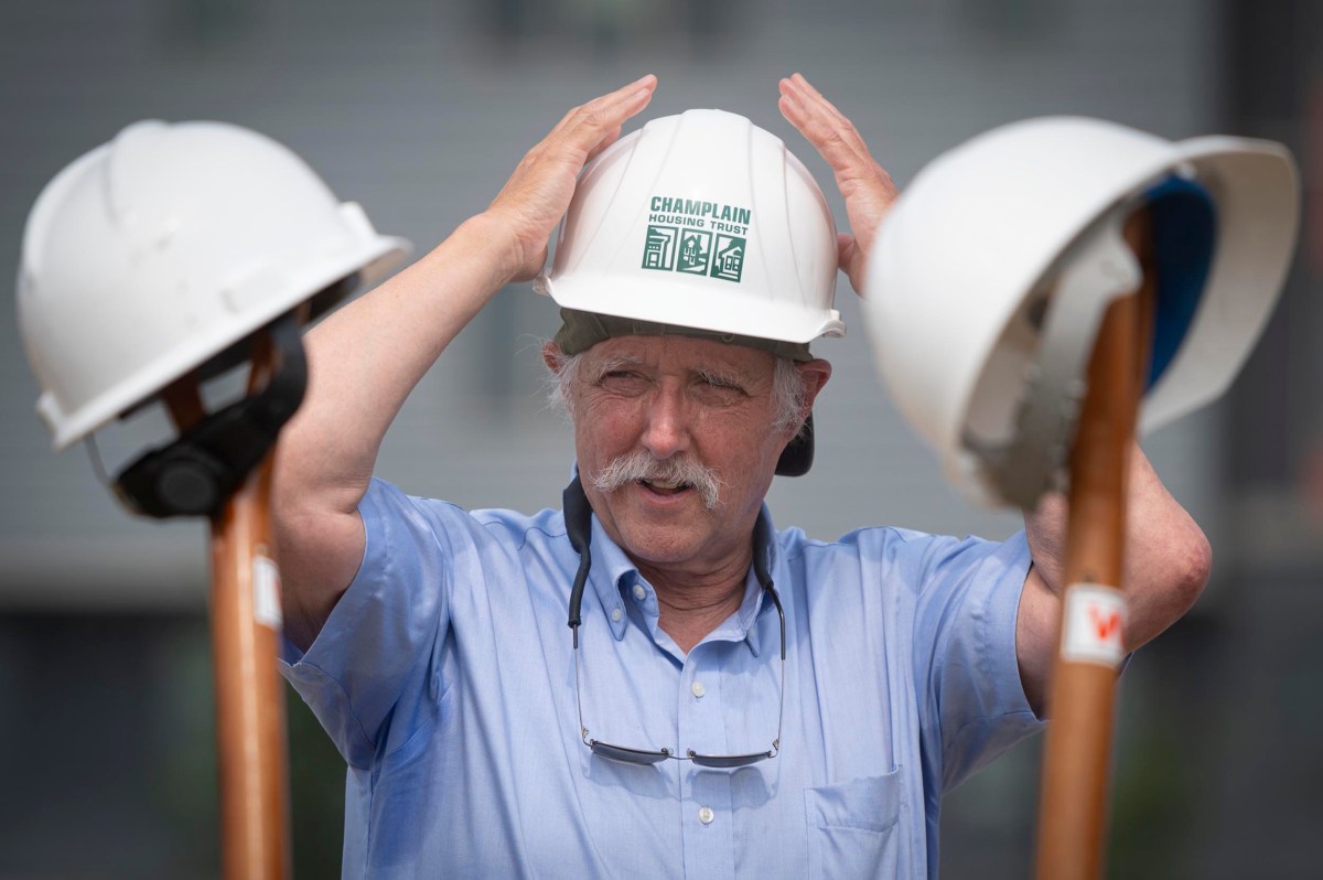 An older man in a blue shirt adjusts a hard hat labeled "CHAMPLAIN," with two other hard hats resting on shovels in the foreground.
