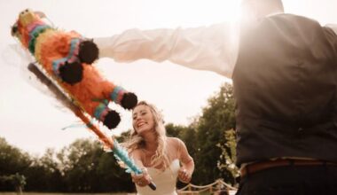 A smiling bride swings a bat at a colorful piñata held by someone in a vest and dress shirt during an outdoor celebration, with sunlight streaming and trees in the background.