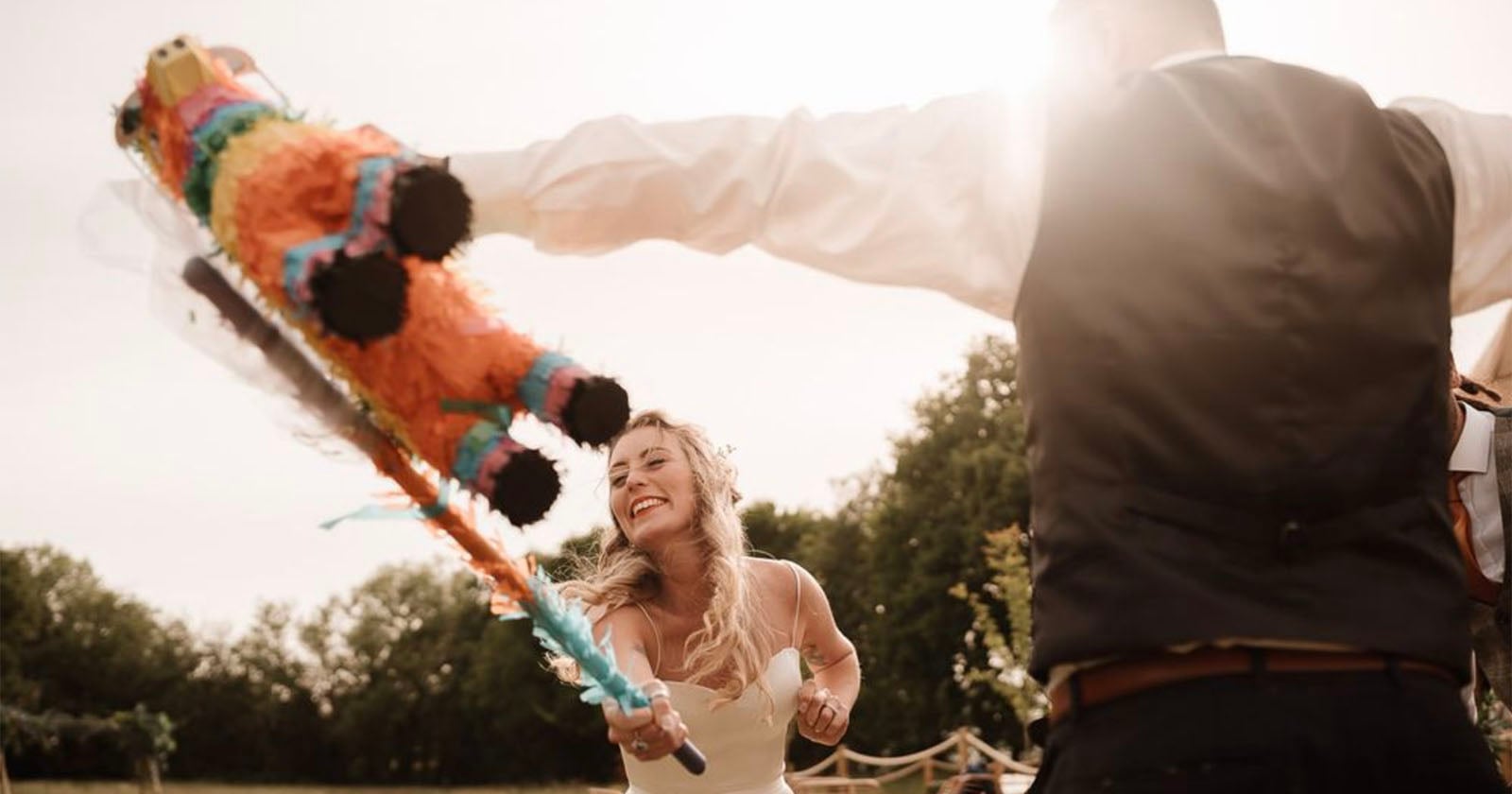 A smiling bride swings a bat at a colorful piñata held by someone in a vest and dress shirt during an outdoor celebration, with sunlight streaming and trees in the background.