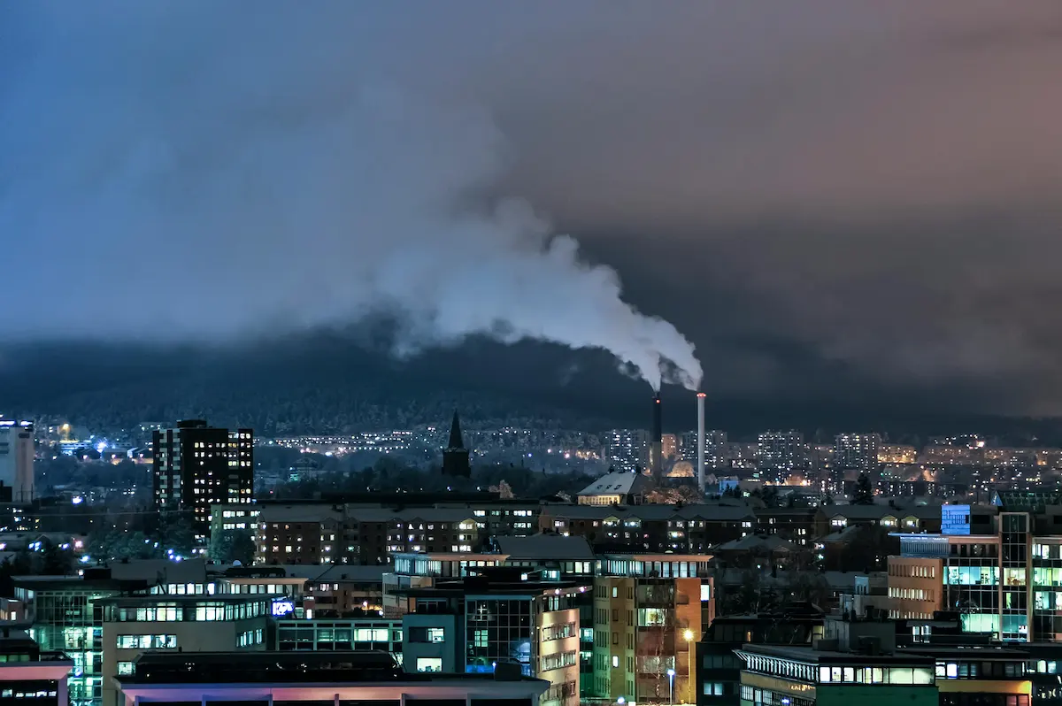 Nighttime city view with many lit buildings. Two industrial chimneys, one black and one white, glow red at their tops while releasing thick white smoke into the dark, cloudy sky.