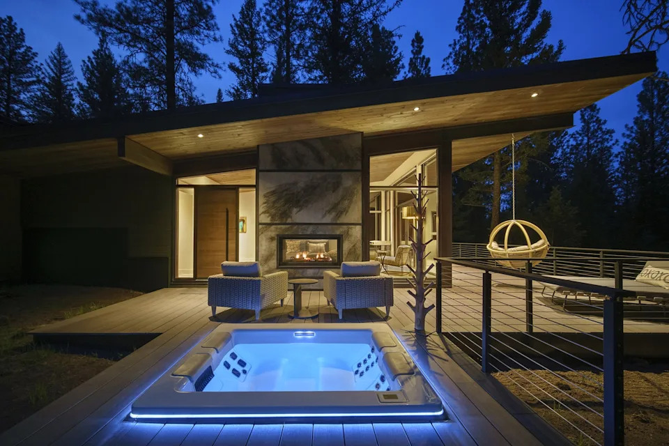 A square hot tub with blue lights in the foreground, set into a deck extending out from a small warmly lit cabin. A darkening sky above and silhouettes of trees behind.