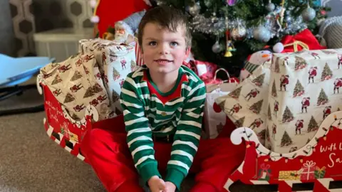 Tammy McDaid A boy with short brown hair is smiling while sitting in front of a Christmas tree. He is dressed in a red, green and white elf costume. There are wrapped presents beneath the tree, along with a red sleigh which says 'with love Santa' on the side.