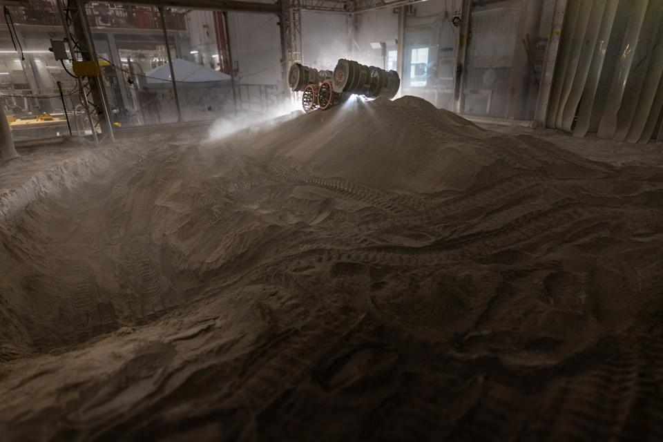 A lunar rover navigating a large indoor dune terrain, kicking up clouds of dust. The dimly lit, industrial setting creates a moody, adventurous atmosphere.