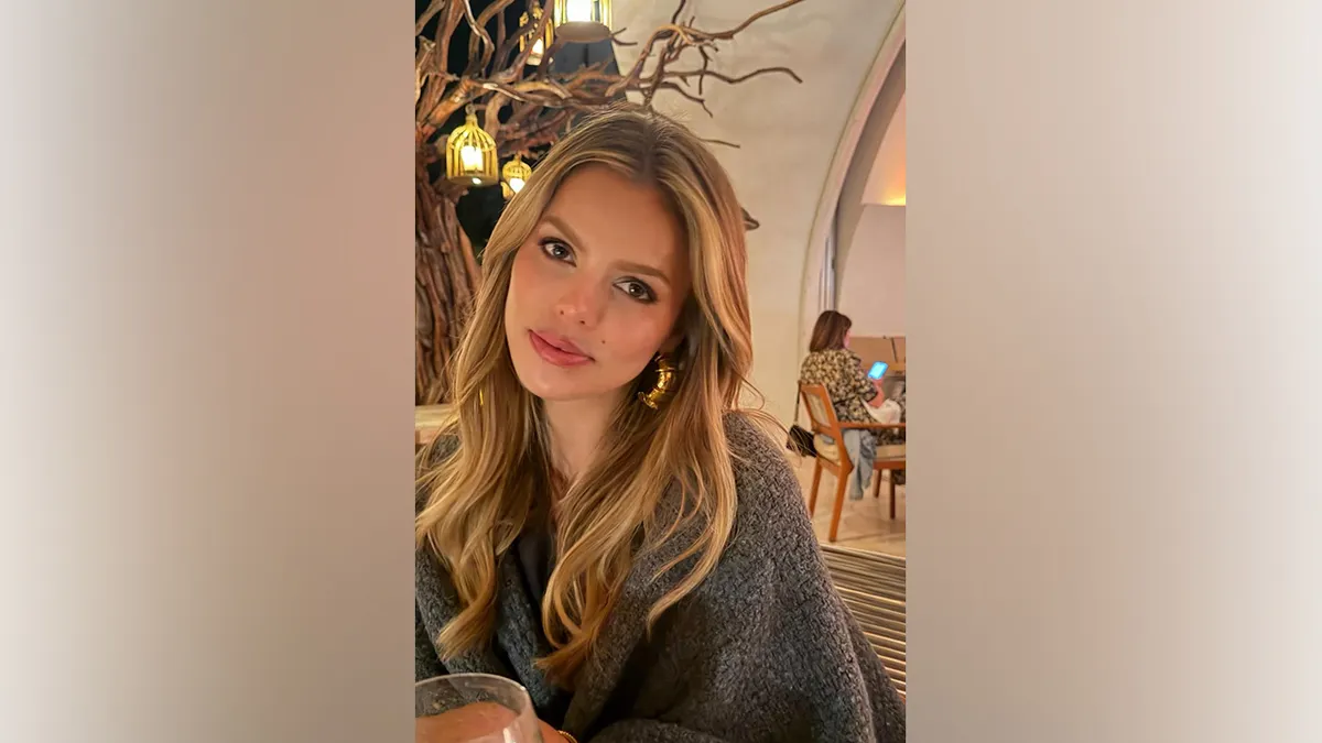 Brooks Nader sits indoors at a restaurant wearing a gray wrap and gold hoop earrings while dining out during her Cabo San Lucas vacation.