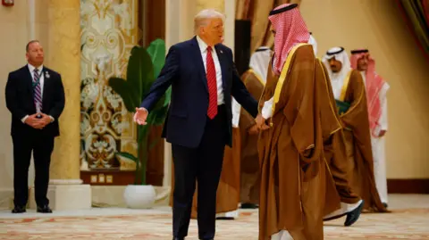 Reuters  Donald Trump gestures next to Saudi Crown Prince Mohammed Bin Salman. Trump wears a dark suit and red tie, while the prince is in a long, brown robe.