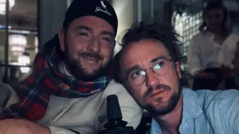 David Holmes David wearing a tartan scarf and a Yankees hat next to Tom who is wearing a blue shirt and glasses. They are inside in a dimly lit area.