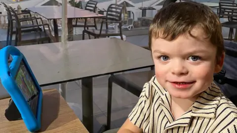 Tammy McDaid A little boy with short brown hair and a blue and cream striped shirt is smiling while seated at a table in a hotel. He has a blue tablet on the table in front of him.