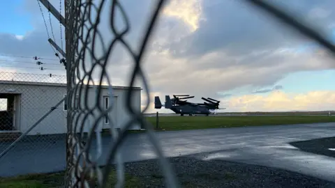 BBC A military aircraft on a runway, photographed through a chain-link fence. 