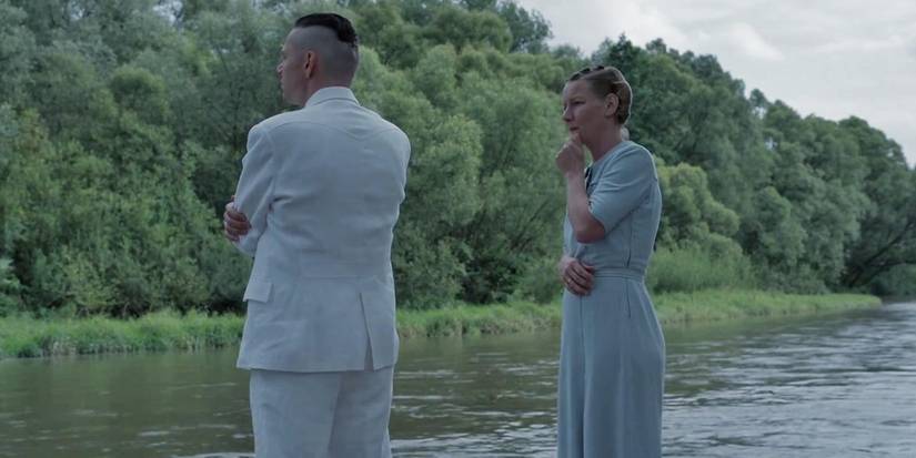 Christian Friedel as Rudolf Höss, Sandra Hüller as Hedwig Höss stand by a river bank in The Zone of Interest.