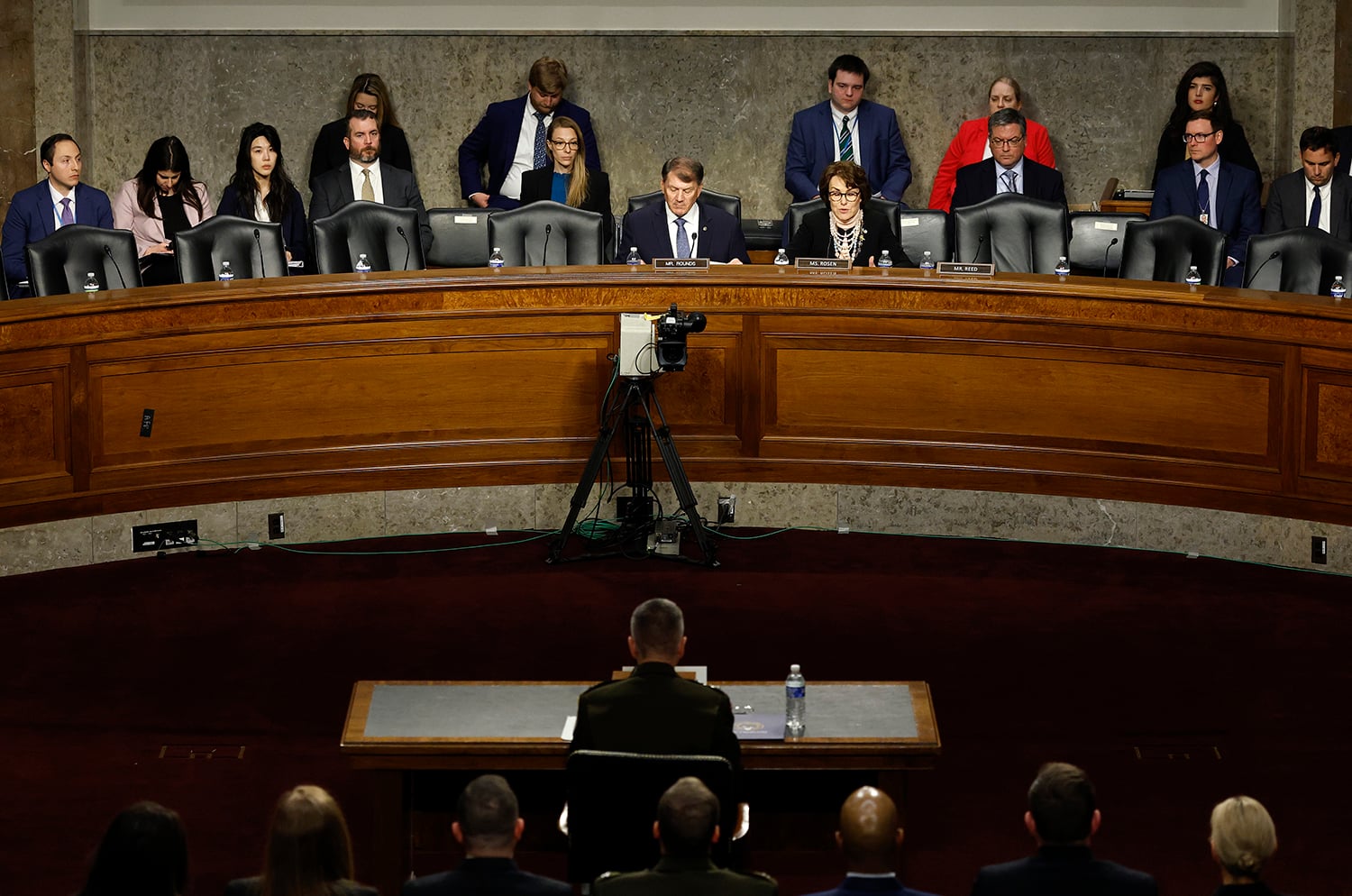 A man is seen from behind at a table of microphones as he testifies before a group of senators on an arched dais above him.