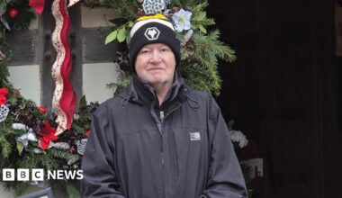 A man in a grey coat and yellow and black bobble hat in front of a wall with green plants and red ribbons