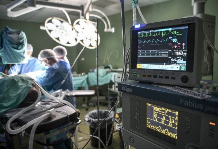 A close-up of a machine showing a person's vitals during surgery, which is being conducted in the background
