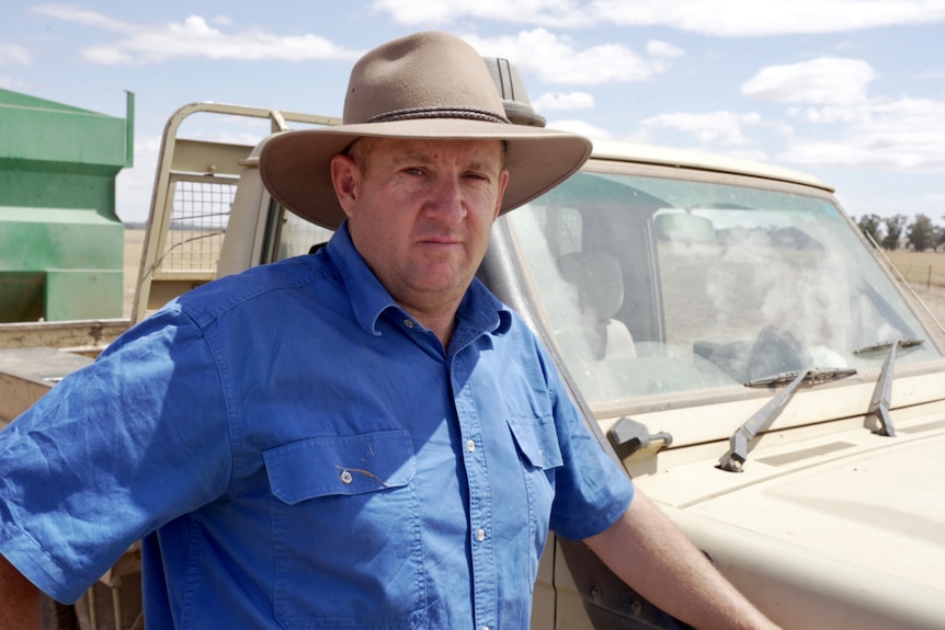 A farmer in a blue shirt and hat next to his ute.