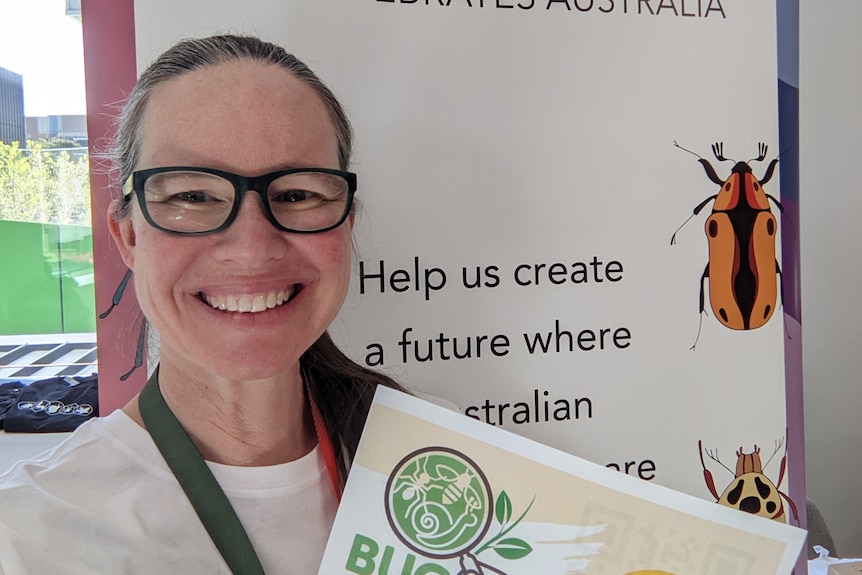 A woman with glasses smiling and holding up a poster with the title 'bug hunt' on it.