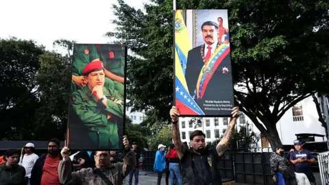 Getty Images Two men hold posters of the late Hugo Chávez (left) and Venezuela's President Nicolás Maduro (right) above their heads in front of large green trees and a small gathering of people