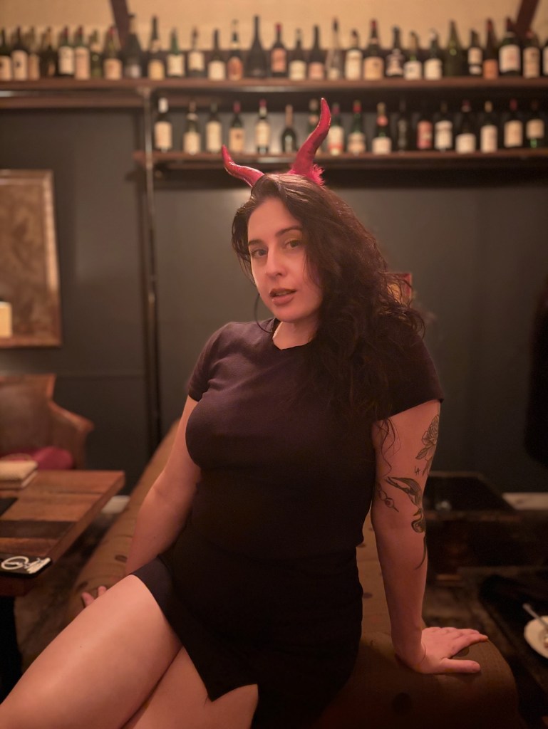 A woman in a black dress with red devil horns sits on a bench in a bar.
