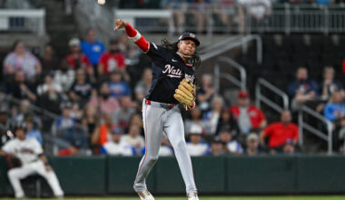 C.J. Abrams, wearing a dark blue jersey and cap with the Nationals team logo, makes a right-handed throw to first base from in-between shortstop and third base.