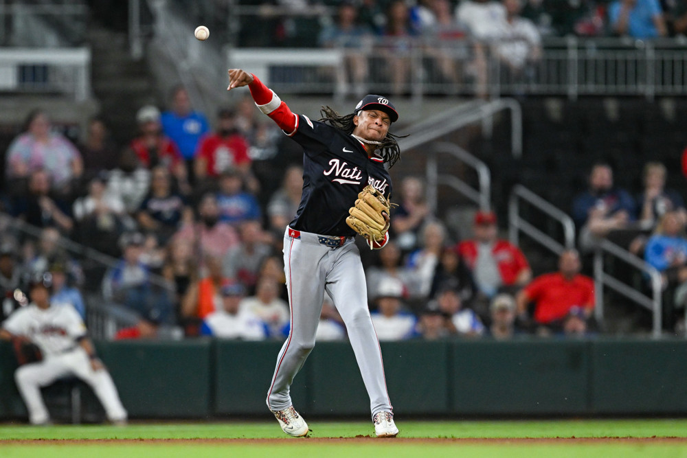 C.J. Abrams, wearing a dark blue jersey and cap with the Nationals team logo, makes a right-handed throw to first base from in-between shortstop and third base.