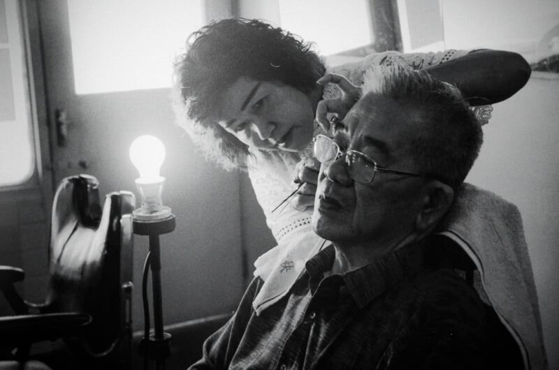 A woman carefully cleans an older man's ear as he sits in a chair, wearing glasses. A lit lightbulb glows nearby, and the scene appears gentle and focused in black and white.