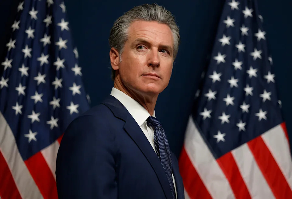 A man in a suit stands between two American flags, glancing to the side. His expression is serious, suggesting a formal event or announcement