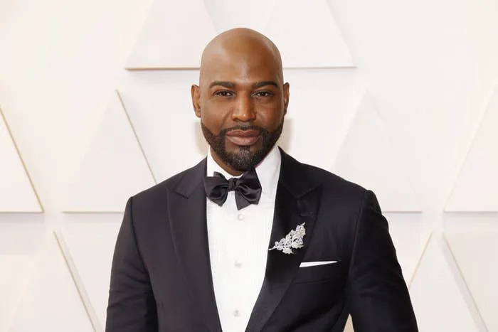 Karamo Brown in a tuxedo with a bow tie and brooch, posing on a geometric-patterned background at a celebrity event