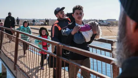 EPA Families cross a narrow bridge as a man carries a small child, with others walking behind him, moving toward safety near a riverside area.