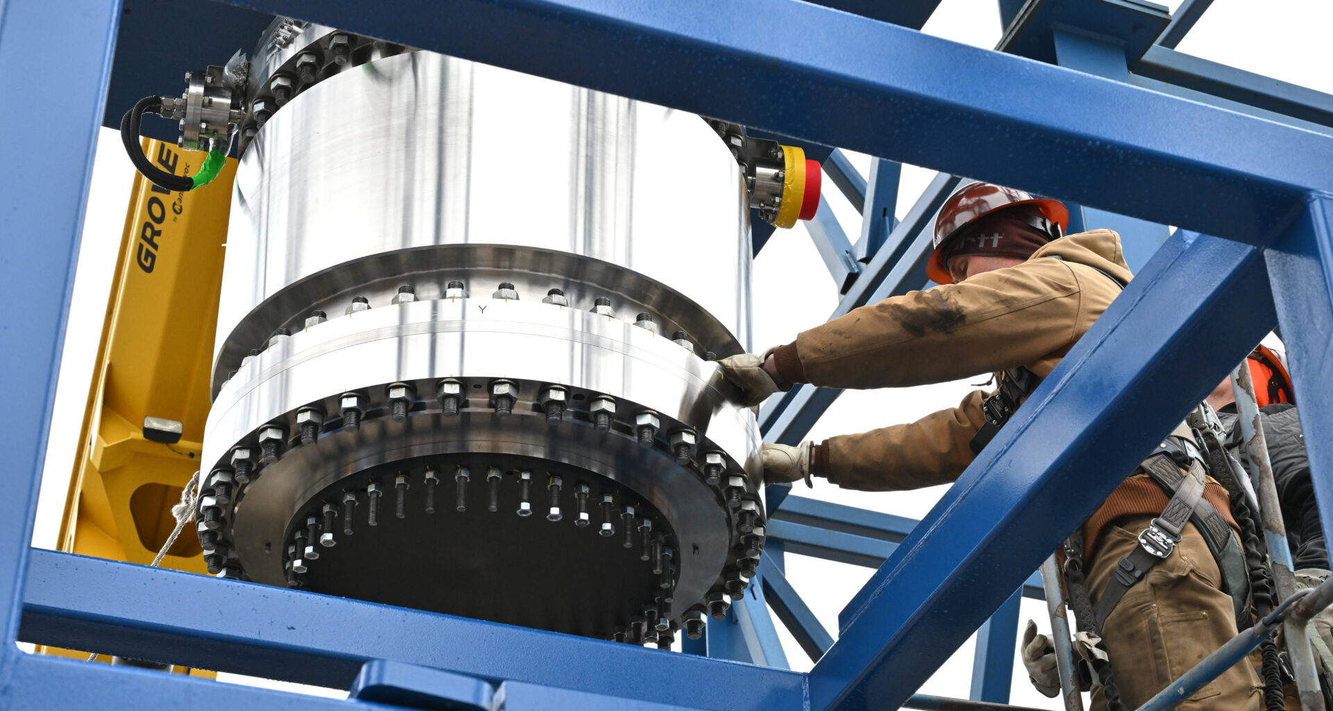 Workers in safety gear guide a large white cylindrical rocket motor or engine component with a black mounting flange as it's positioned within a blue steel test stand structure. Yellow crane equipment is visible on the left side