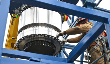 Workers in safety gear guide a large white cylindrical rocket motor or engine component with a black mounting flange as it's positioned within a blue steel test stand structure. Yellow crane equipment is visible on the left side