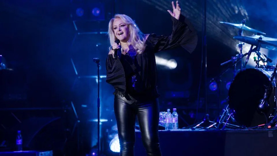 Bonnie Tyler dressed in black clothes, on stage and singing. One hand holds a microphone, the other is raised in the air above her head. The stage is light by blue lighting, and a drum kit can be seen behind her. 