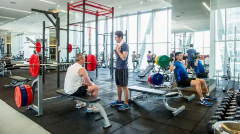 Getty Images People training in what looks like a commercial gym. One man is sat on a weight bench chatting to a man stood opposite him. In the right corner, a man is lifting dumbells with both arms. In the background, women can be seen using the bike machines. 