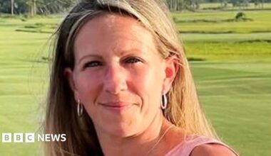 A woman wearing a pink shirt holds a glass while posing in front of a green lawn.