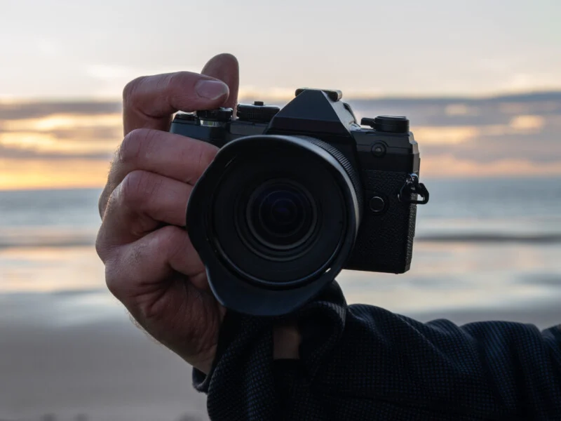 A close-up of a hand holding a black camera, with the lens pointing toward the viewer. In the background, there is a blurred beach scene at sunset, with soft light and clouds in the sky.