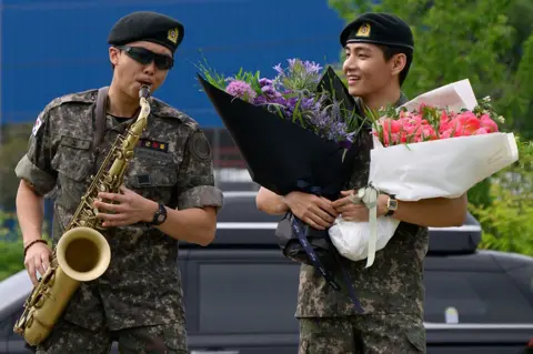 Getty Images K-pop boy band BTS members RM (L) plays the saxophone while V looks on while holding flower bouquets at an outdoor sporting facility in Chuncheon on June 10, 2025