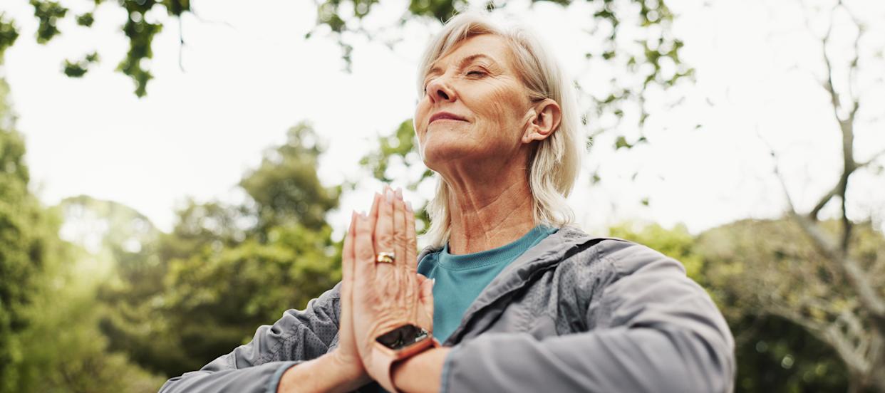 Close up of an older woman outside with her hands pressed together and eyes closed, meditating or doing yoga.