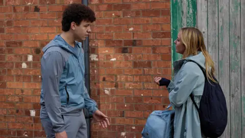 ITV A TV still of Beth Nixon stood opposite the character of Will Driscoll. Nixon is wearing a rain coat and carrying a bag. Driscoll is wearing a light blue and grey tracksuit. 