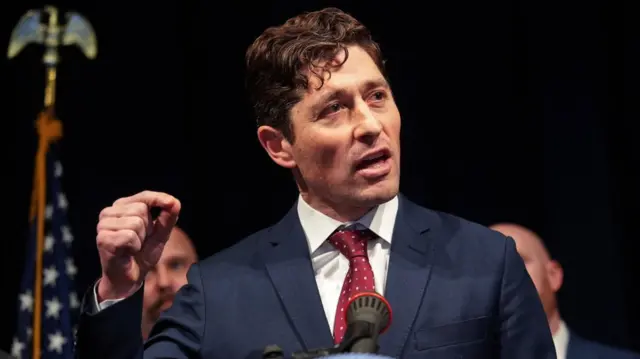 Minneapolis mayor Jacob Frey pictured mid-speech with his right hand raised. He's wearing a dark blue suit, white shirt and polka-dotted red tie, two men partially visible behind him. A US flag attached to a pole stands to Frey's right in the background