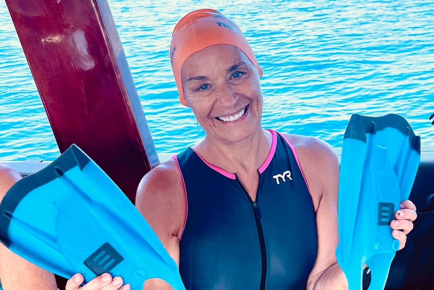 A woman in a swimsuit and a swimming caps smiles holding two flippers while sitting on a boat.